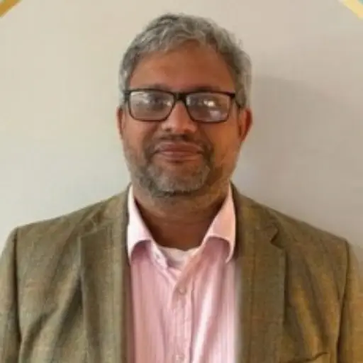 Man with gray hair and beard wearing glasses, pink shirt and brown blazer, smiling at the camera.
