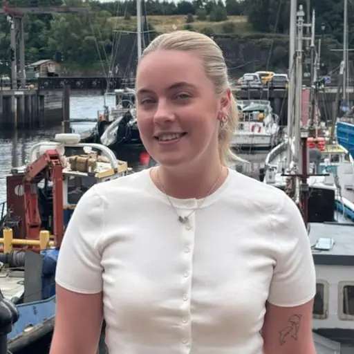 Young woman with blonde hair tied back in a white short-sleeve buttoned top, standing at a marina with boats and water in the background.